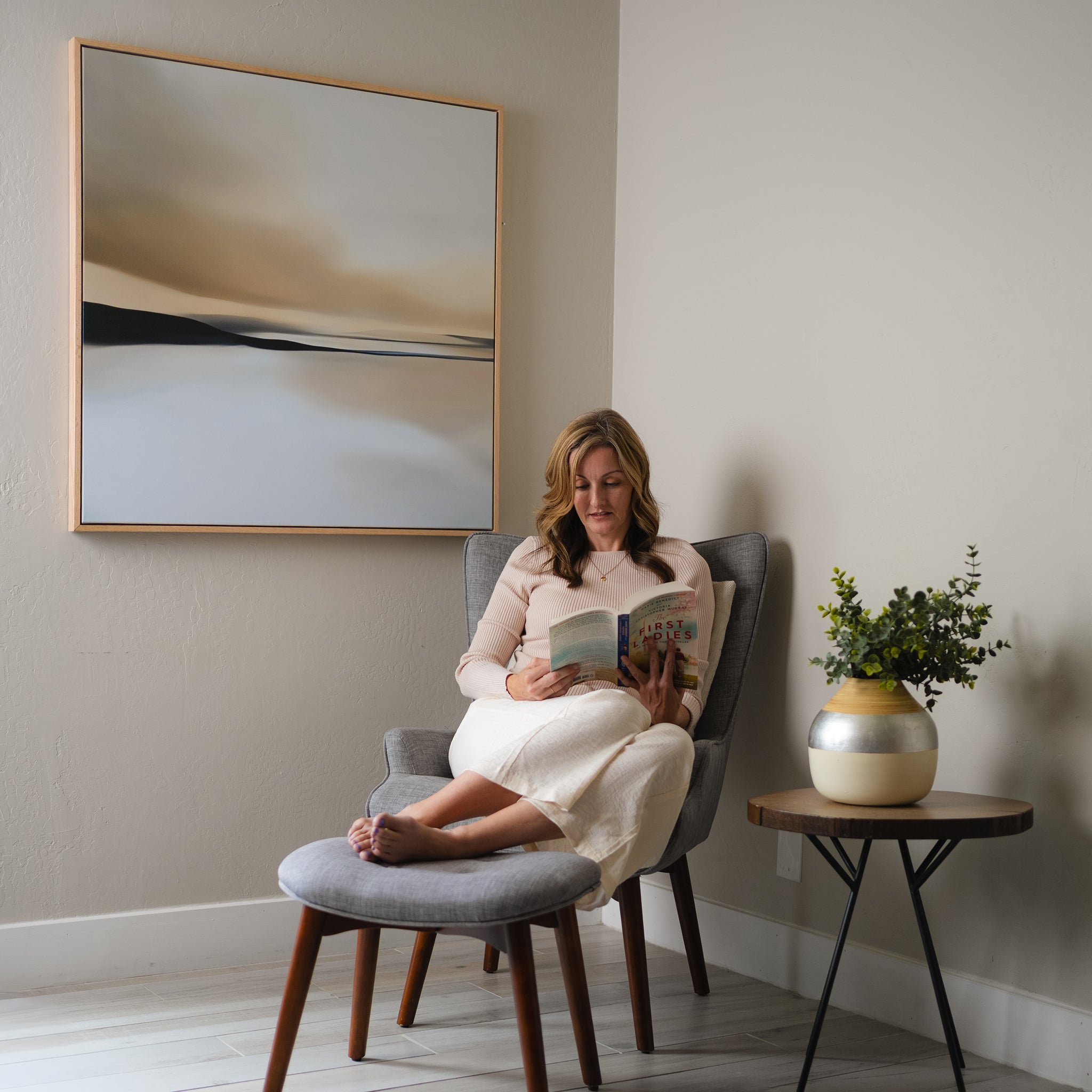 A woman reads in a gray armchair with an ottoman in a minimalist living room. The Desert Quietude Abstract Canvas Art hangs on the wall, while a round side table with a vase of greenery adds to the decor.
