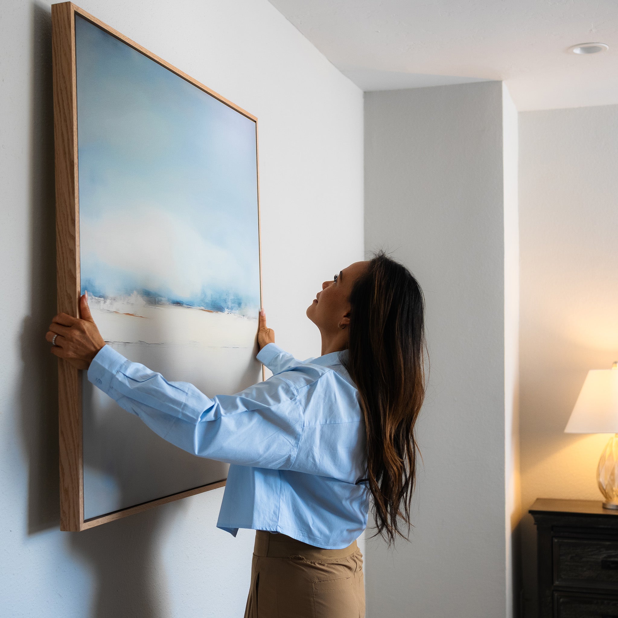 A woman with long dark hair hangs the Misty Morning Abstract Canvas Art, a minimalist wall painting, on a white wall in a bright room. A lamp on a dark wooden table appears in the background, enhancing the tranquil atmosphere.