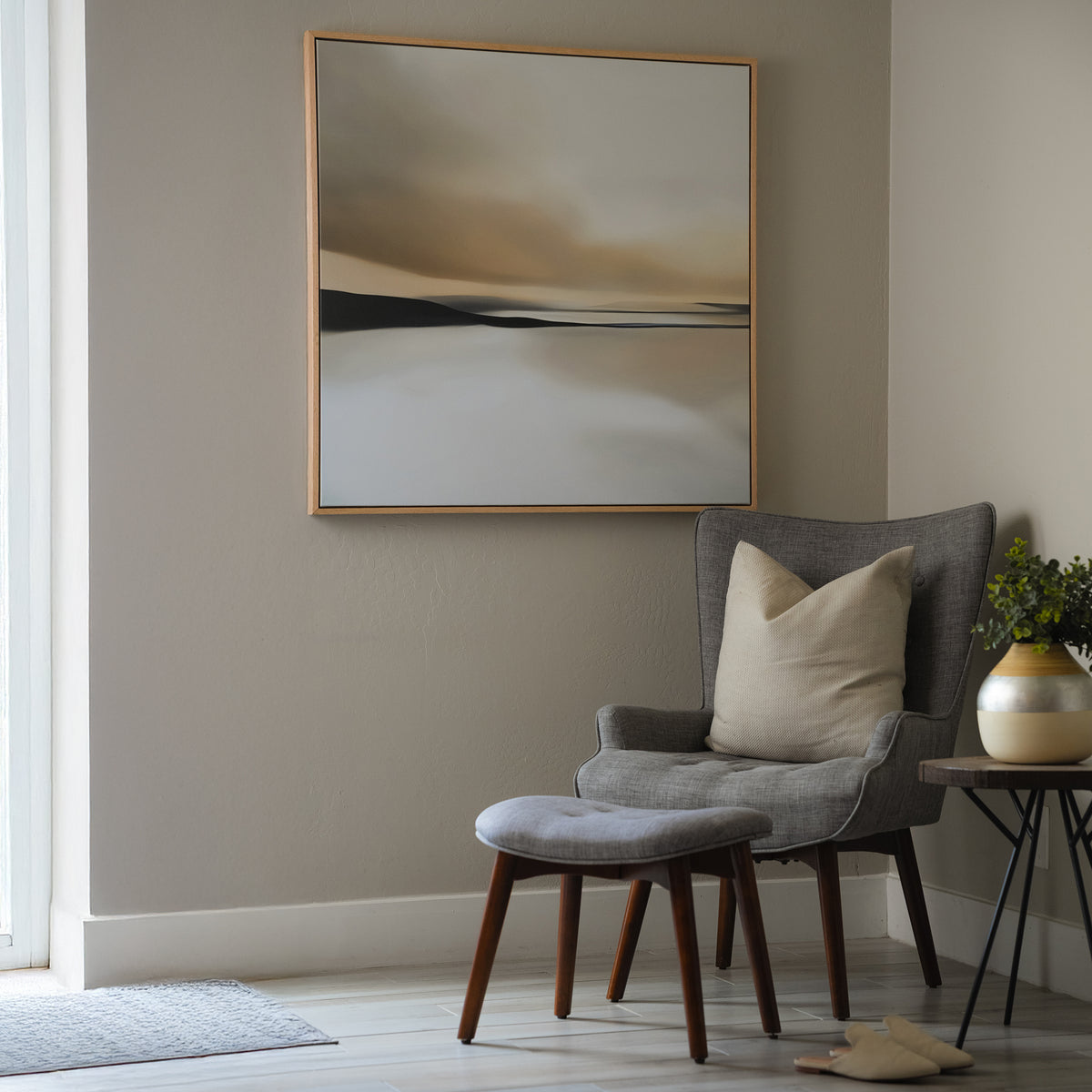 A gray armchair with ottoman and beige pillow forms a cozy corner. Nearby, a side table holds flowers, and the Desert Quietude Abstract Canvas Art hangs above on the neutral wall, as sunlight brightens the inviting living room.