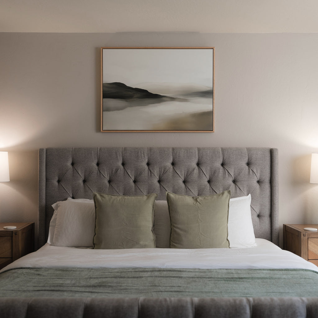 A neatly made bed with a tufted gray headboard, three green pillows, and white bedding sits between two bedside tables with lamps. Above the bed, Soft Valley Canvas Art – Horizontal enhances the minimalist vibe of the light-colored wall.