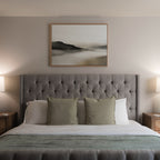 A neatly made bed with a tufted gray headboard, three green pillows, and white bedding sits between two bedside tables with lamps. Above the bed, Soft Valley Canvas Art – Horizontal enhances the minimalist vibe of the light-colored wall.
