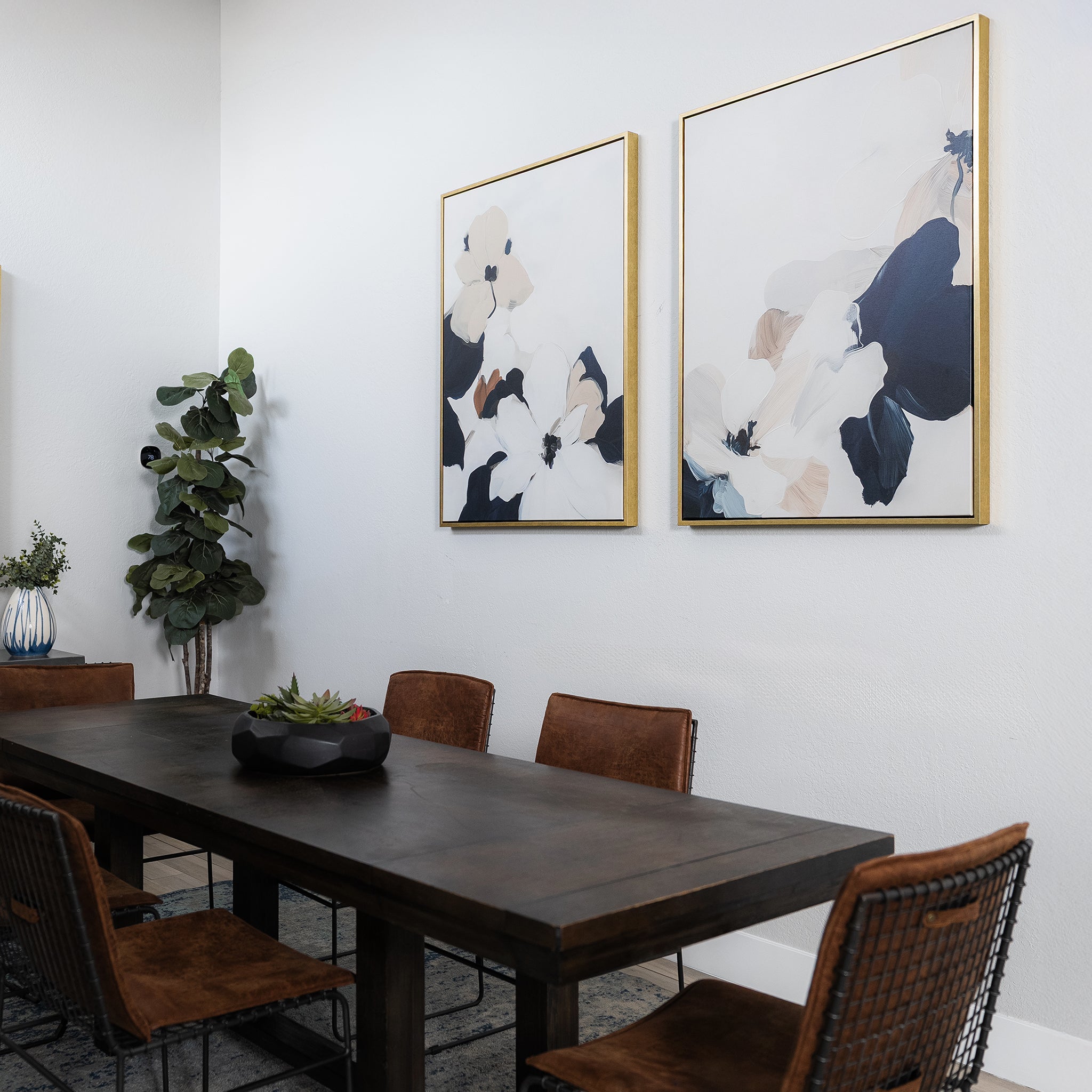 A modern dining room with a dark wooden table, six brown chairs, a potted plant in the corner, the Floral Serenity Set of 2 minimalist wall art on the wall, and a dark centerpiece on the table.