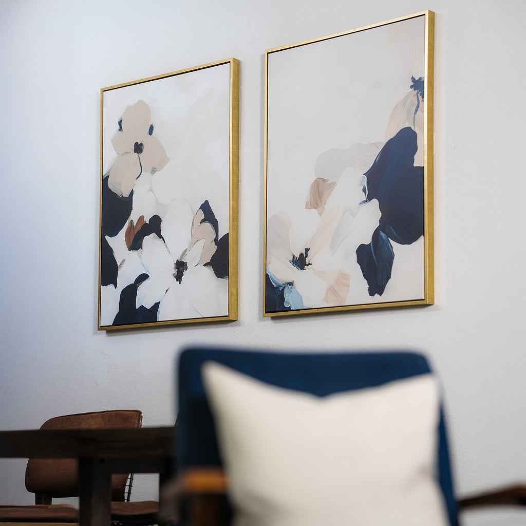 The Floral Serenity Set of 2, featuring abstract floral art in gold frames, hangs on a light wall. A blurred cushion and part of a wooden table and chair in the foreground enhance this minimalist living room decor setup.