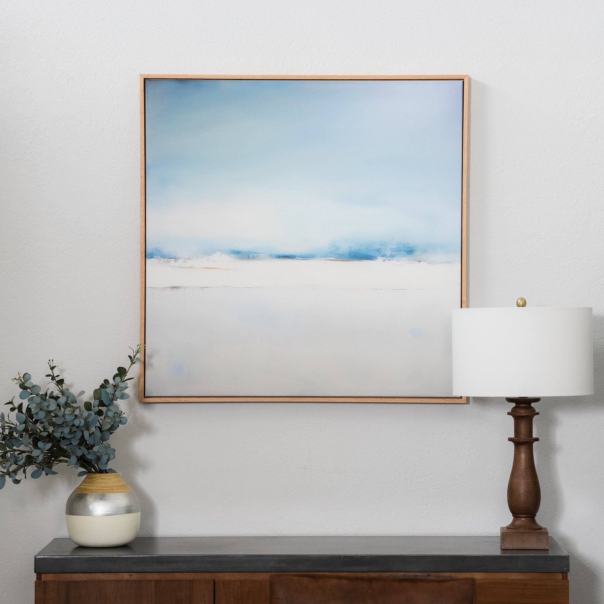 The Misty Morning Abstract Canvas Art, with soft blue and white tones, hangs above a wooden sideboard. Ideal for coastal decor, it adds tranquility when paired with a vase of greenery and a table lamp with a wooden base.