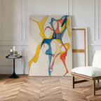 The Dance Of Unity Abstract Canvas Art, featuring flowing yellow, blue, and red shapes, leans against a white paneled wall beside an empty frame, a round table with candles, and a cushioned chair on chevron wood flooring.