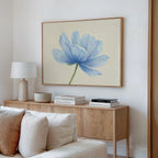 A modern living room features a wooden sideboard with books, decor, and a white table lamp. Above the white sofa hangs the Daybreak Flower Canvas Art—a large minimalist print of a blue flower.