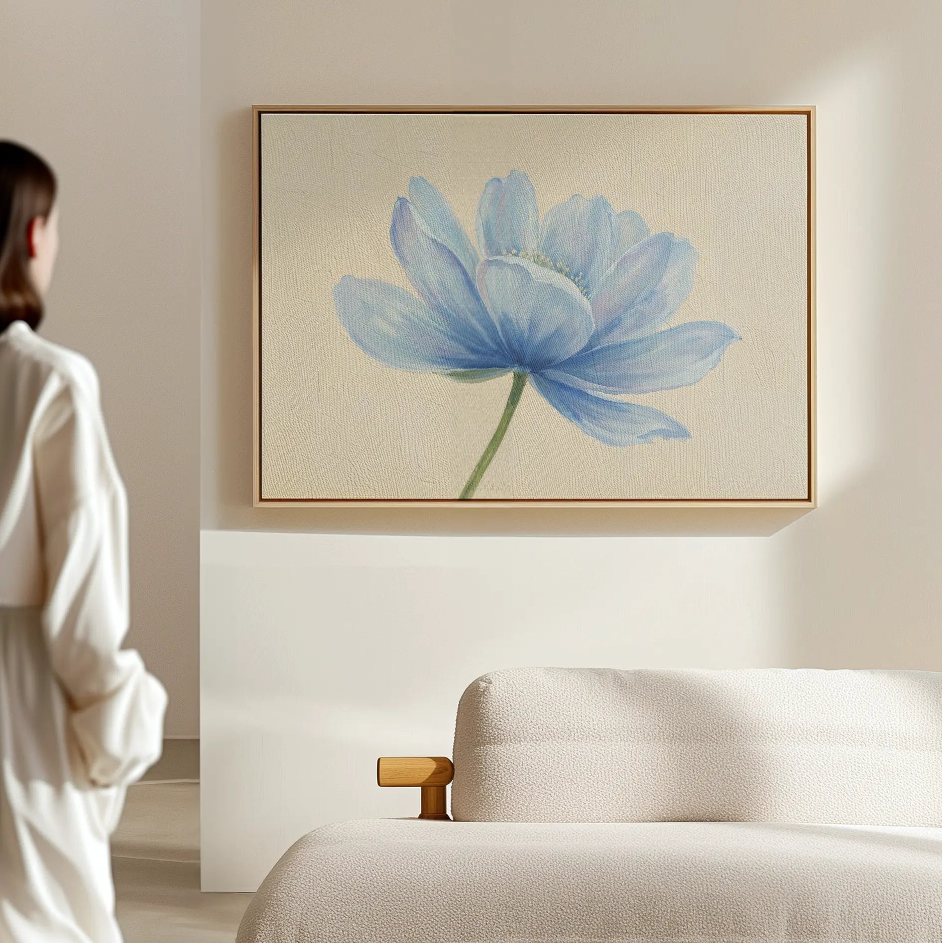 A person stands in a minimal, light-filled room with a textured sofa and the Daybreak Flower Canvas Art, a large modern wall piece featuring a blue flower—ideal for bedroom decor.