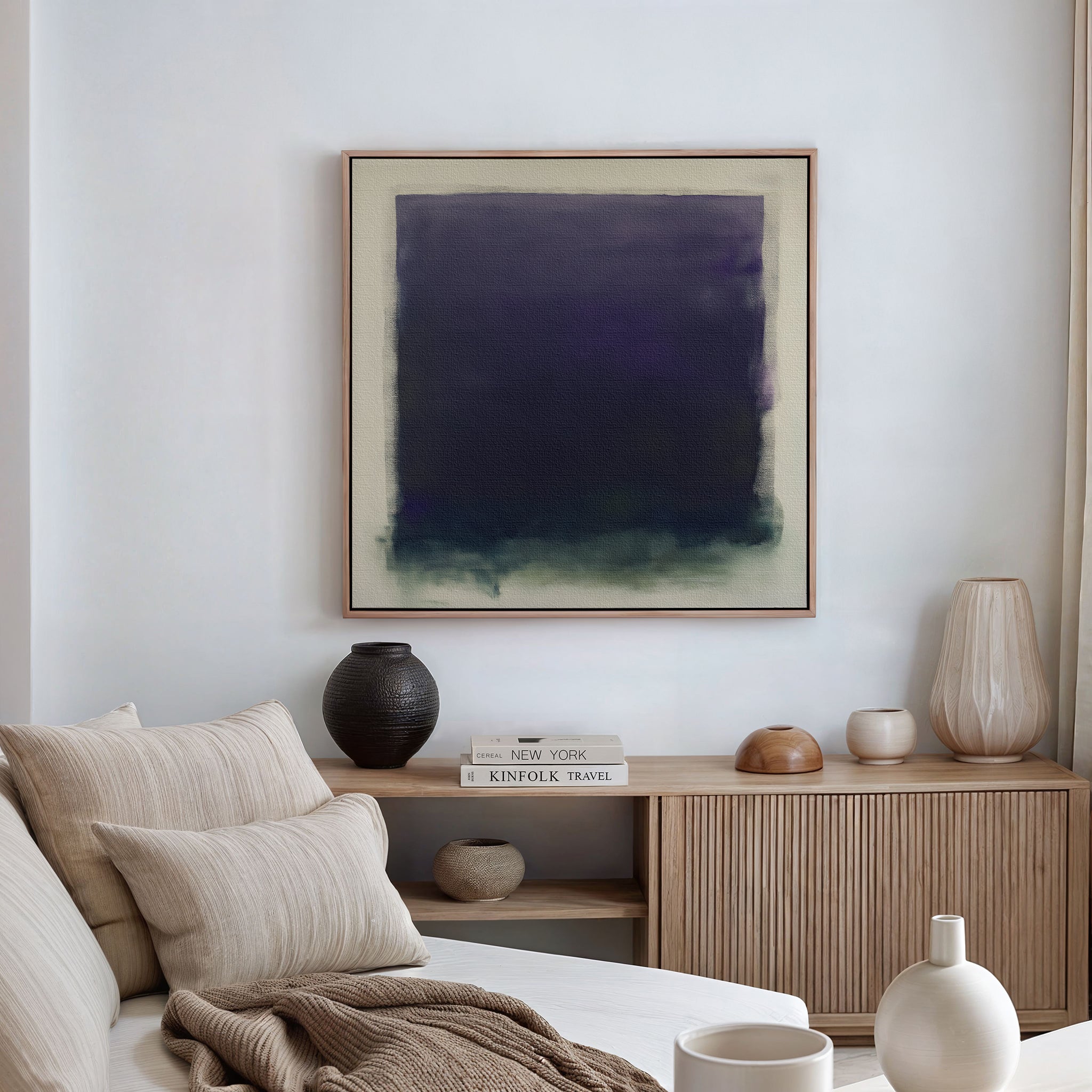 A modern living room features a beige sofa with neutral cushions, a wooden sideboard with books and vases, and the striking Deepest Night Canvas Art in dark blue and green as minimalist wall decor.