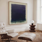 A modern, minimalist room features a white leather lounge chair with ottoman, a wooden side table with a white dome lamp, and the Deepest Night Canvas Art in dark blue and green tones displayed prominently on the wall.
