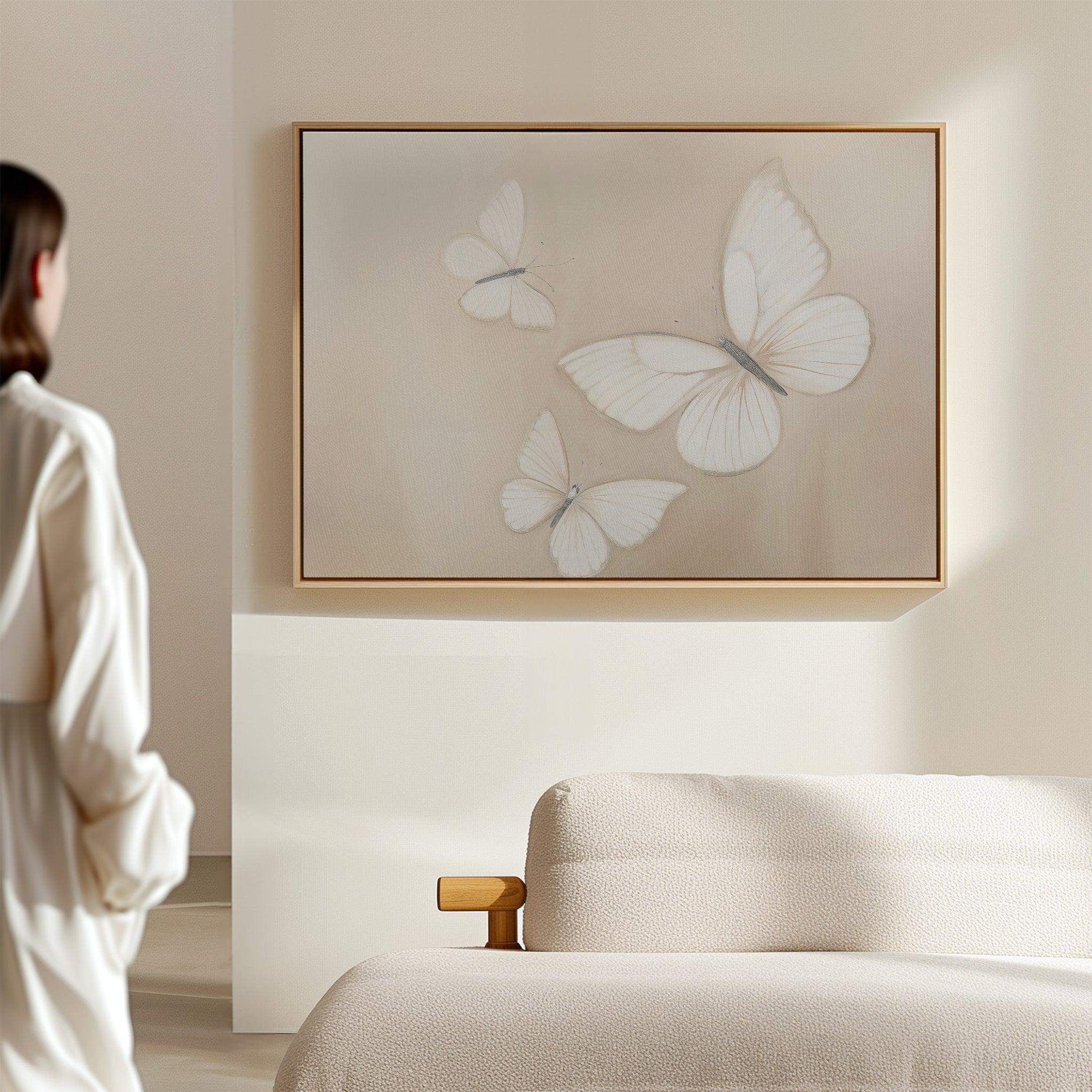 A person admires Delicate Whites Canvas Art—three white butterflies framed—on a beige wall above a light sofa, adding elegant neutral decor to this softly lit, Nordic Park-inspired room.