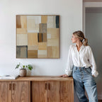 A woman in a white blouse and jeans leans against a wooden cabinet, admiring the Desert Mosaic Abstract Canvas Art on the wall. A small potted plant and a book on the cabinet enhance the cozy living room decor.