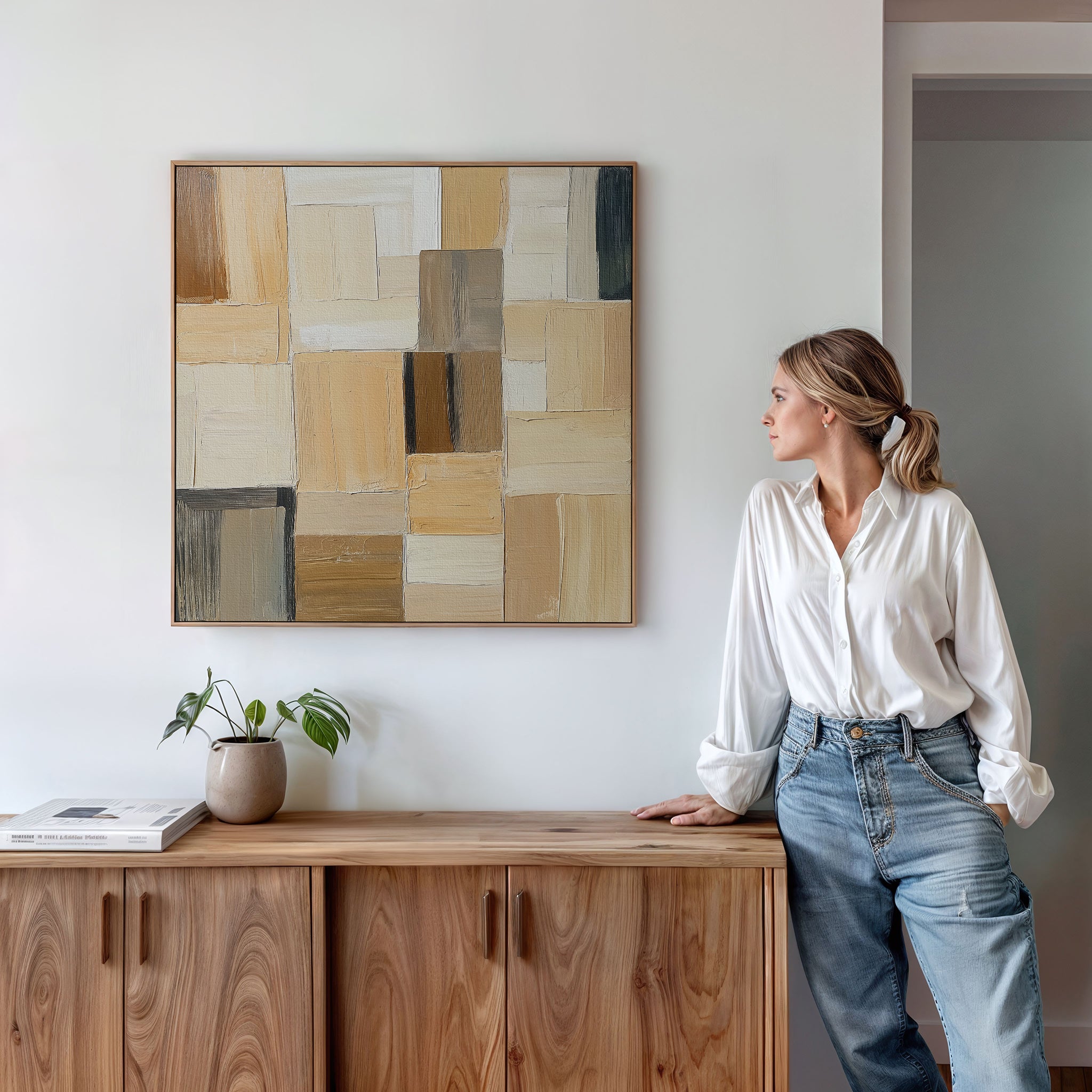 A woman in a white blouse and jeans leans against a wooden cabinet, admiring the Desert Mosaic Abstract Canvas Art on the wall. A small potted plant and a book on the cabinet enhance the cozy living room decor.