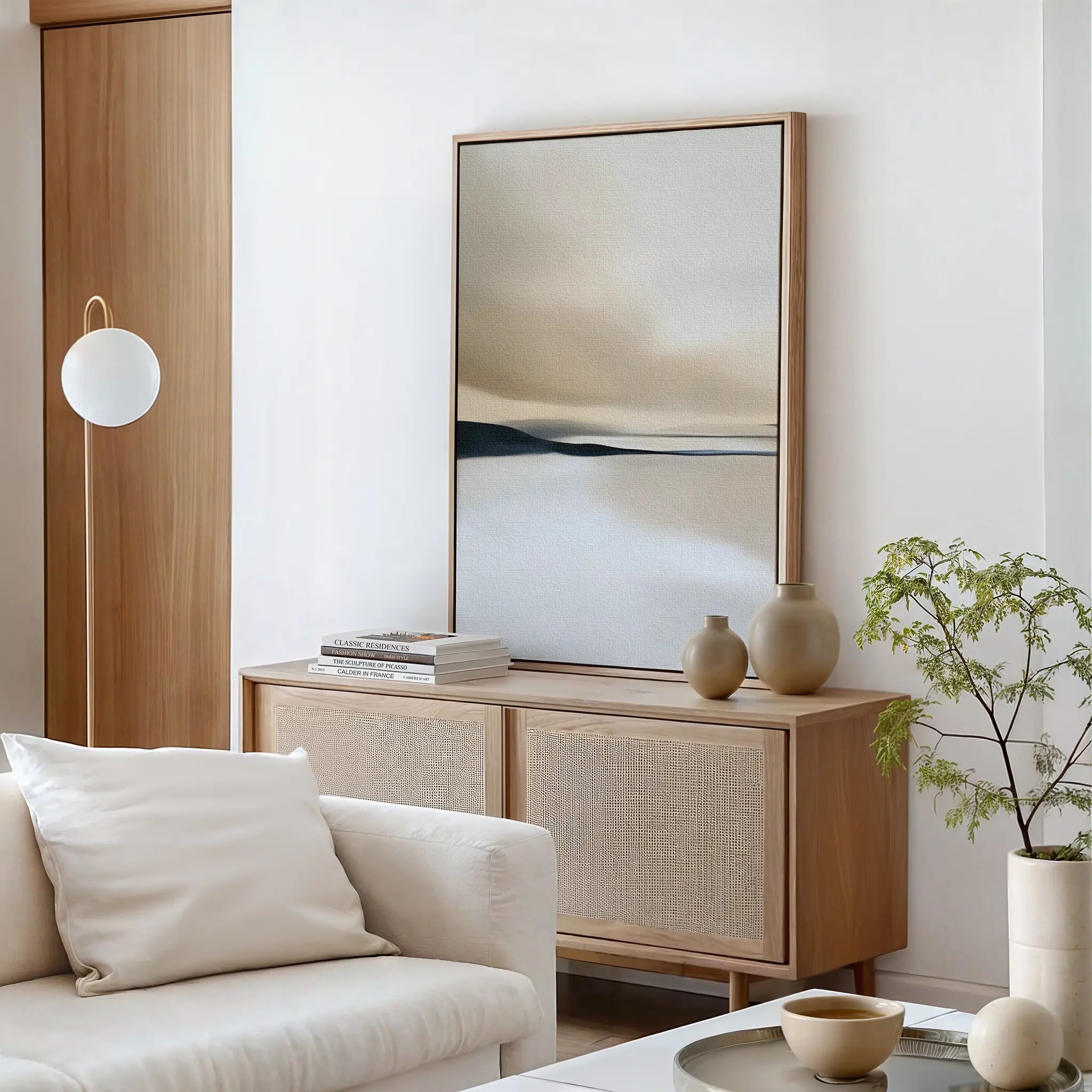 A minimalist living room features a light wooden sideboard, the Desert Quietude Canvas Art – Vertical, ceramic vases, stacked books, a white sofa with a pillow, a round lamp, and a small green plant on a glass coffee table.