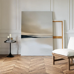 A minimalist living space with herringbone wood floors showcases the Desert Quietude Canvas Art – Vertical against a white-paneled wall. A small round table with candles and books and a modern chair complete the stylish decor.