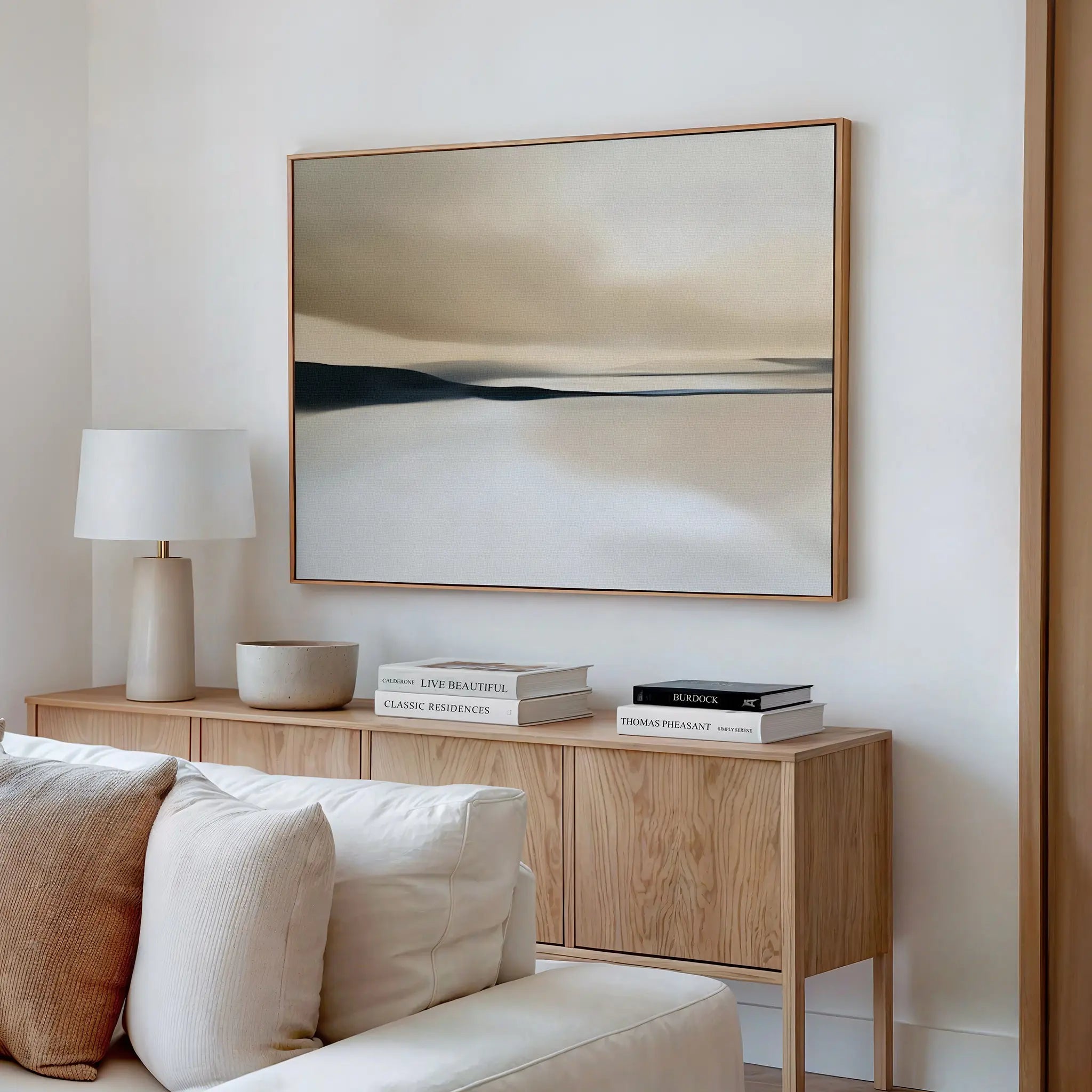A minimalist living room featuring a light wood sideboard, stacked books, a white lamp, a bowl, and the Desert Quietude Canvas Art – Horizontal in neutral tones above a beige sofa.