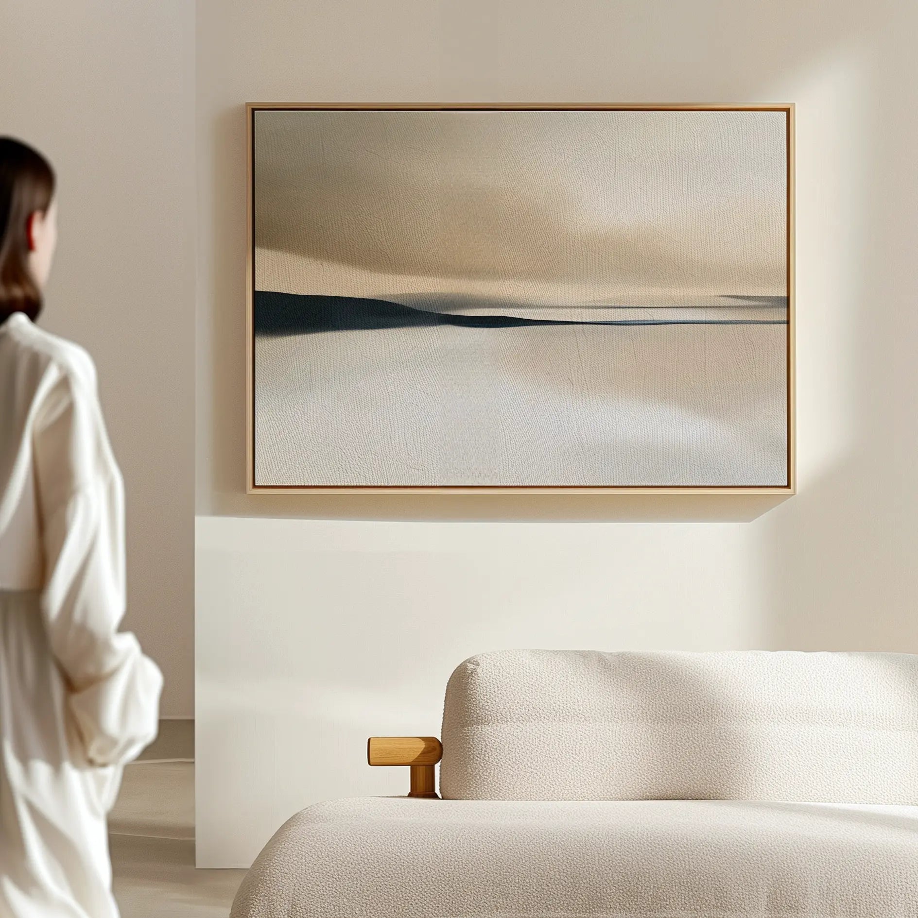 A person in light attire stands in a minimal room, admiring the Desert Quietude Canvas Art – Horizontal in soft beige, white, and blue above a light couch—an ideal choice for modern living room decor.