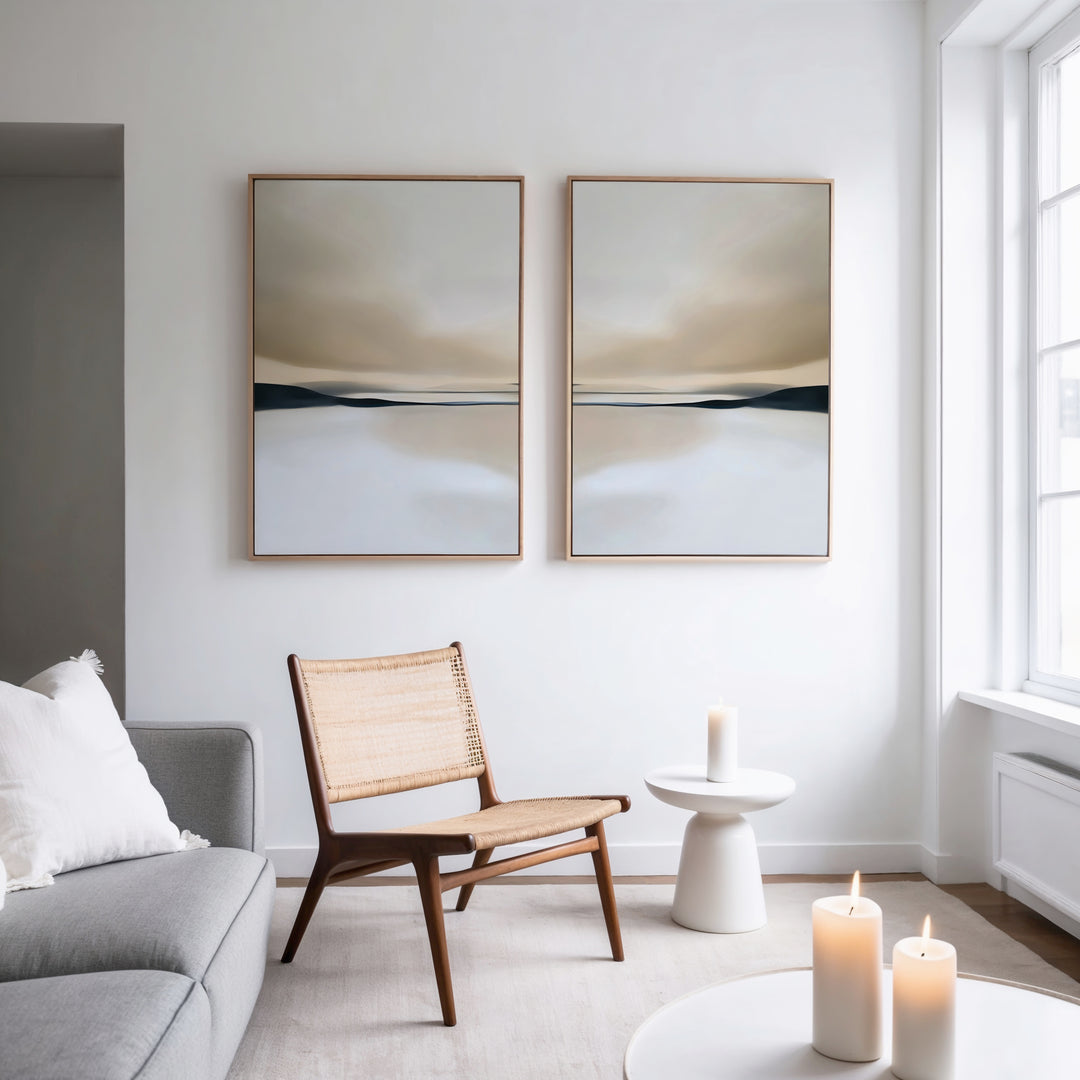 A minimalist living room with a wooden chair, small white table, lit candles, a gray sofa with a white pillow, and the Desert Quietude Set of 2 paintings above. Natural light streams through a large window, highlighting the modern decor.