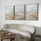 A modern living room features a curved beige sofa, round wooden coffee table with books and flowers, and the Desert Serenity Set of 3 minimalist wall art depicting soft sandy dunes as elegant decor above.