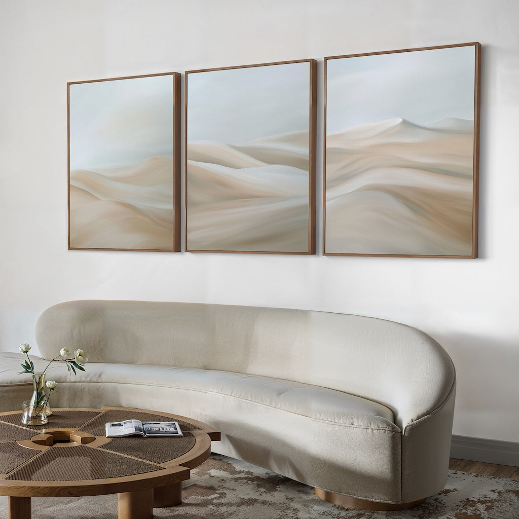 A modern living room features a curved beige sofa, round wooden coffee table with books and flowers, and the Desert Serenity Set of 3 minimalist wall art depicting soft sandy dunes as elegant decor above.