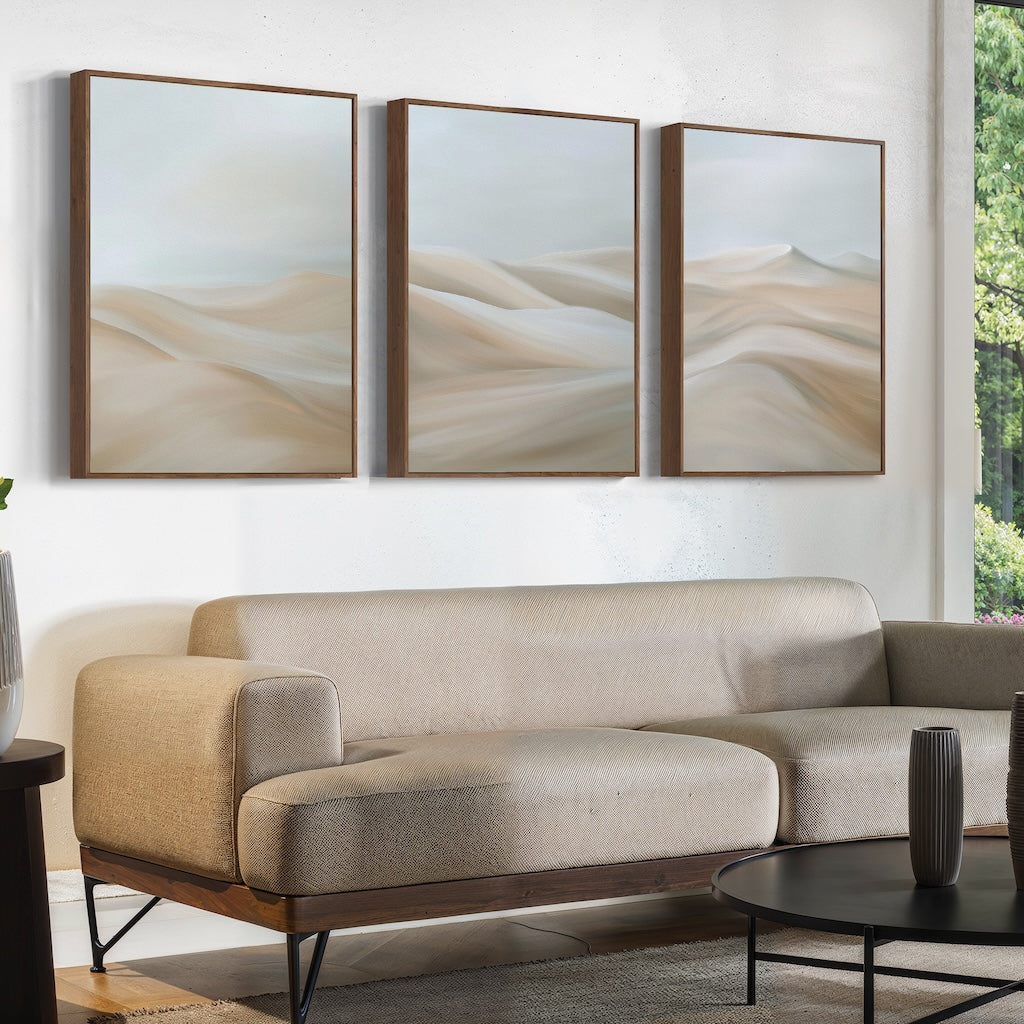 A modern living room with a beige sofa, a round black coffee table, and the Desert Serenity Set of 3 abstract landscape paintings in soft neutrals hanging above the couch. A large window frames lush greenery, enhancing the serene decor.
