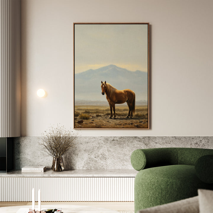 A modern living room in neutral tones showcases the Desert Watch Canvas Art—a large framed painting of a horse in a desert—mounted above a marble shelf with dried flowers, books, and a green curved sofa in the foreground.