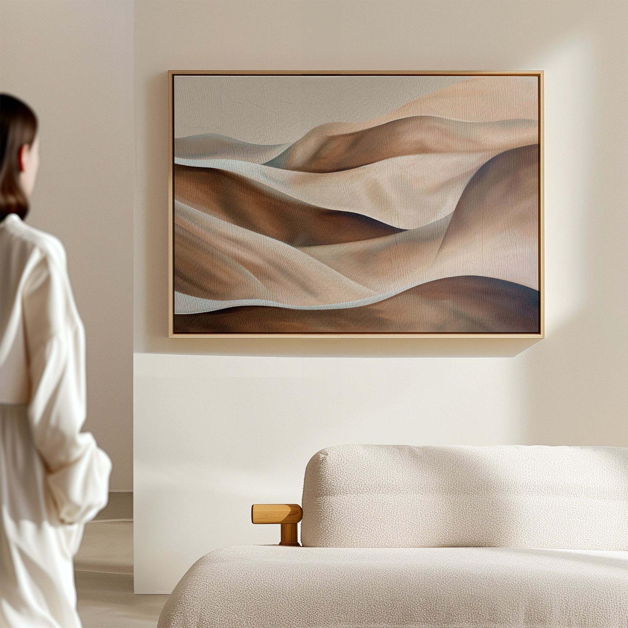 A person in white stands in a minimalist room, admiring the Desert Waves Abstract Canvas Art—a large painting of soft, earthy hills—hanging above a beige curved sofa, adding modern elegance to the space.