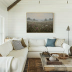 A cozy living room with white shiplap walls, a large white sectional sofa with assorted pillows, a rustic wooden coffee table, and the Distant Fields Canvas Art—a framed landscape painting—hanging above the sofa.