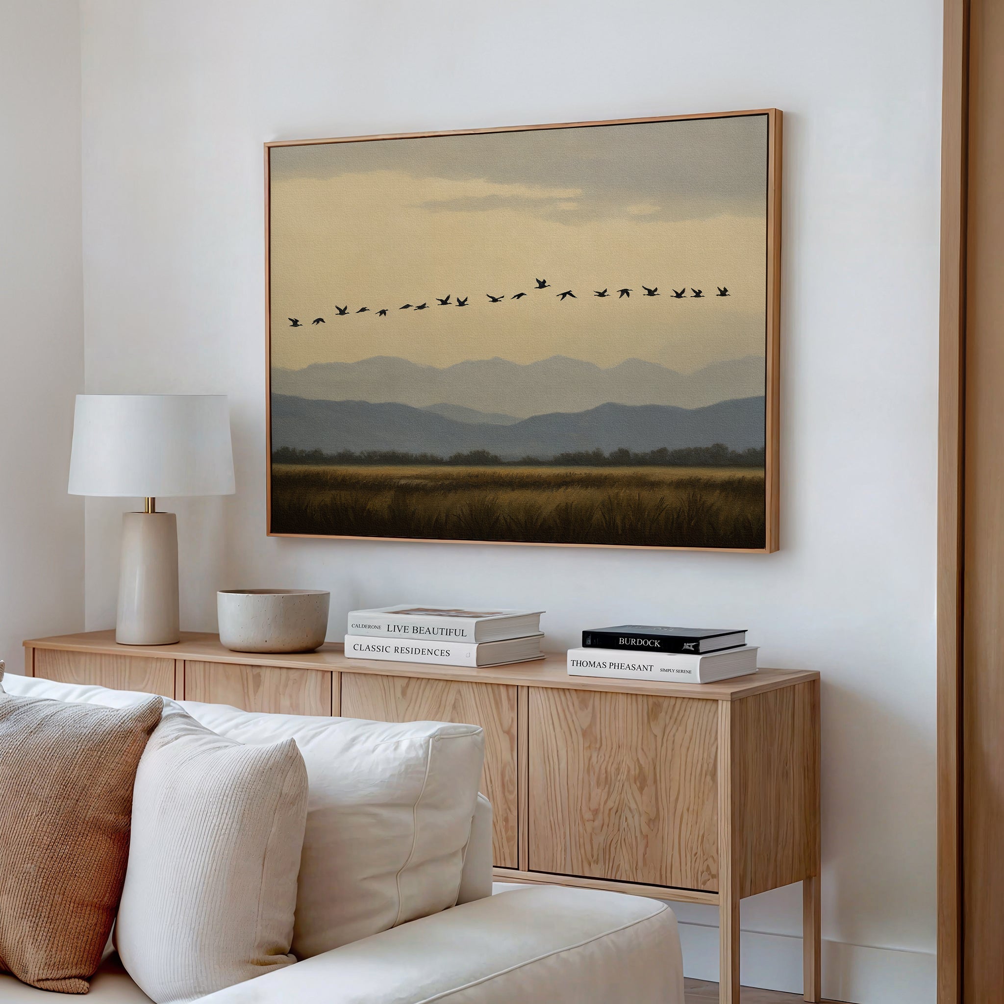 A cozy living room features a light wood sideboard with a white lamp and stacked books, topped by the Distant Flight Canvas Art depicting birds soaring above mountains. A white sofa sits in the foreground.