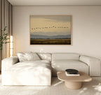 A modern living room features a light beige sectional, wooden coffee table, and the Distant Flight Canvas Art on the wall. Natural light enhances the minimalist decor and the tranquil scene of birds flying over mountains.
