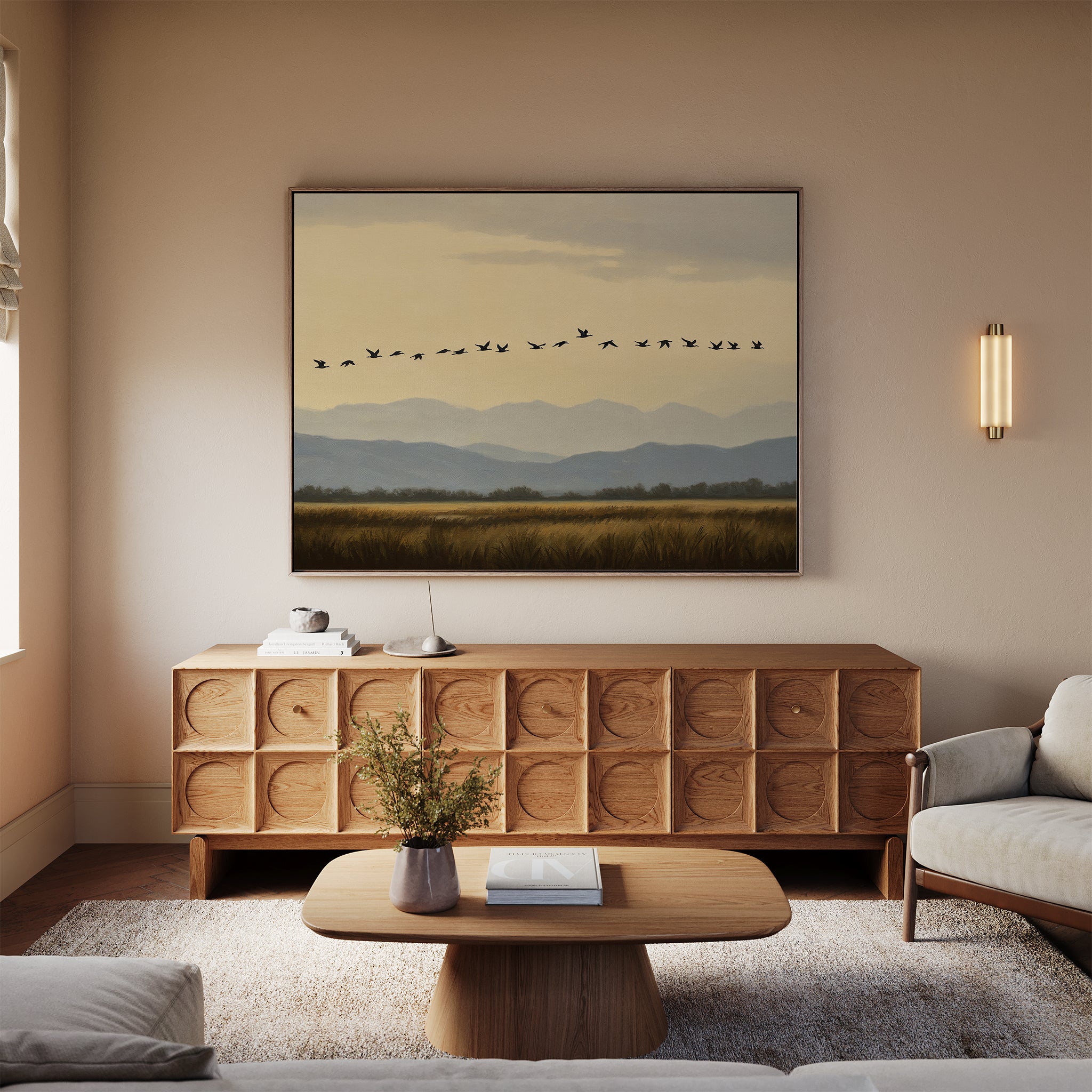 A cozy living room with neutral tones includes a wooden sideboard, low coffee table with a vase of flowers, sofa, and a striking Distant Flight Canvas Art print depicting birds flying over mountains and fields.
