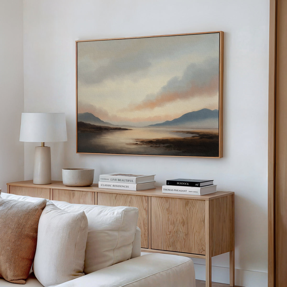 A cozy living room with a light wood sideboard, neutral decor, books, a white lamp, and minimalist wall art—a large Distant Peaks Canvas Art—hanging above the furniture on a white wall.