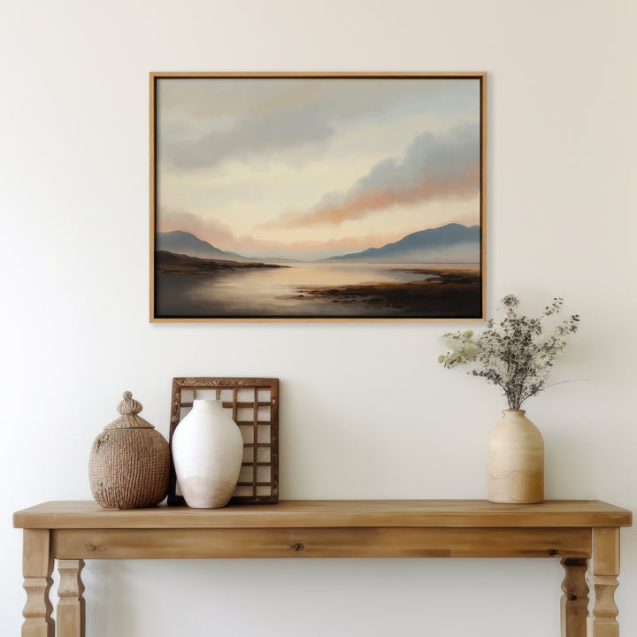 A wooden console table with decorative vases, a wicker basket, and dried flowers sits against a white wall, topped with the Distant Peaks Canvas Art—landscape wall decor of a peaceful lake and mountains under cloudy skies—ideal for living rooms.