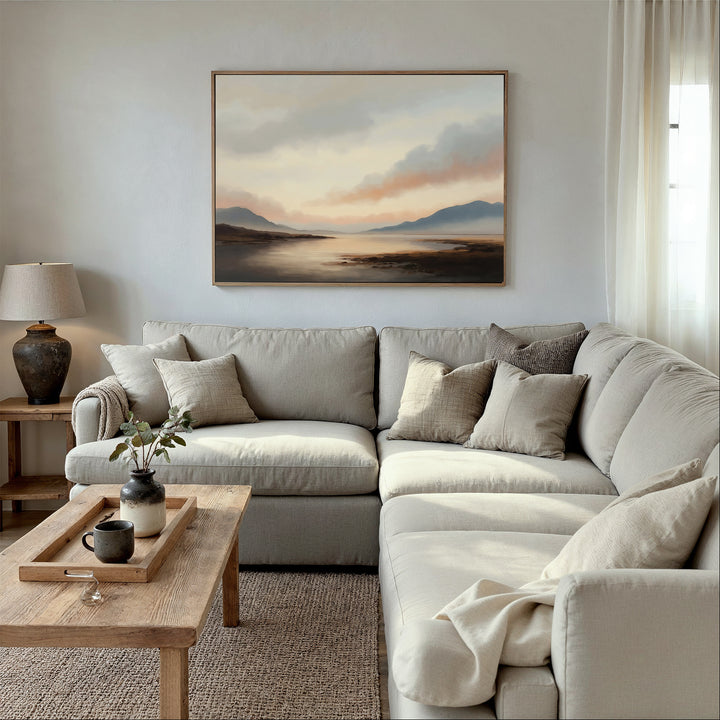 A cozy living room features a beige sectional sofa, neutral throw pillows, a light wooden coffee table with decor, a table lamp, and Distant Peaks Canvas Art above the sofa. Soft natural light enhances the inviting space.