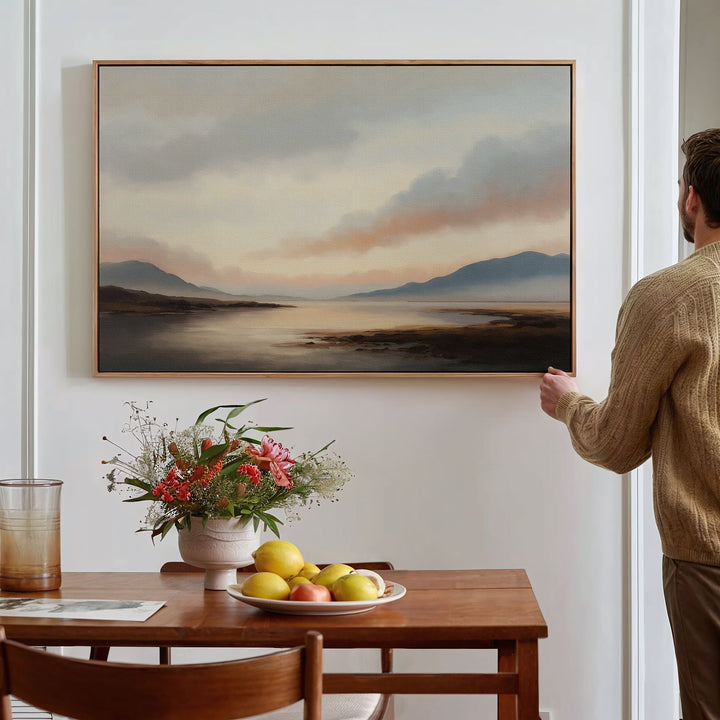 A person hangs the Distant Peaks Canvas Art—a minimalist landscape of mountains and water—on a white wall above a wooden dining table with flowers, fruit, and a glass of juice.