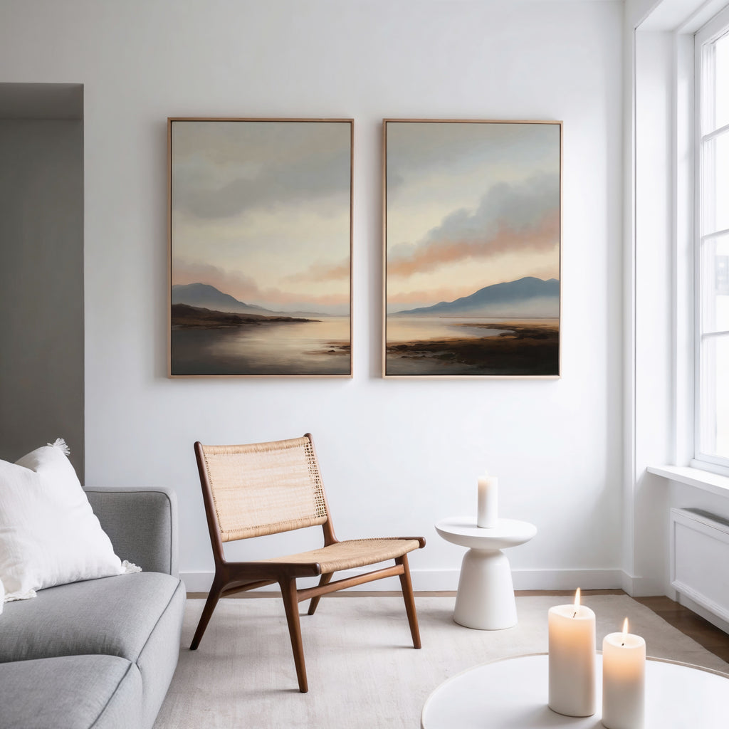 A modern living room features a gray sofa, wooden chair, round white side table, and lit candles. Above, the Distant Peaks Set of 2 minimalist wall art hangs on a white wall near large windows.