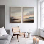 A modern living room features a gray sofa, wooden chair, round white side table, and lit candles. Above, the Distant Peaks Set of 2 minimalist wall art hangs on a white wall near large windows.