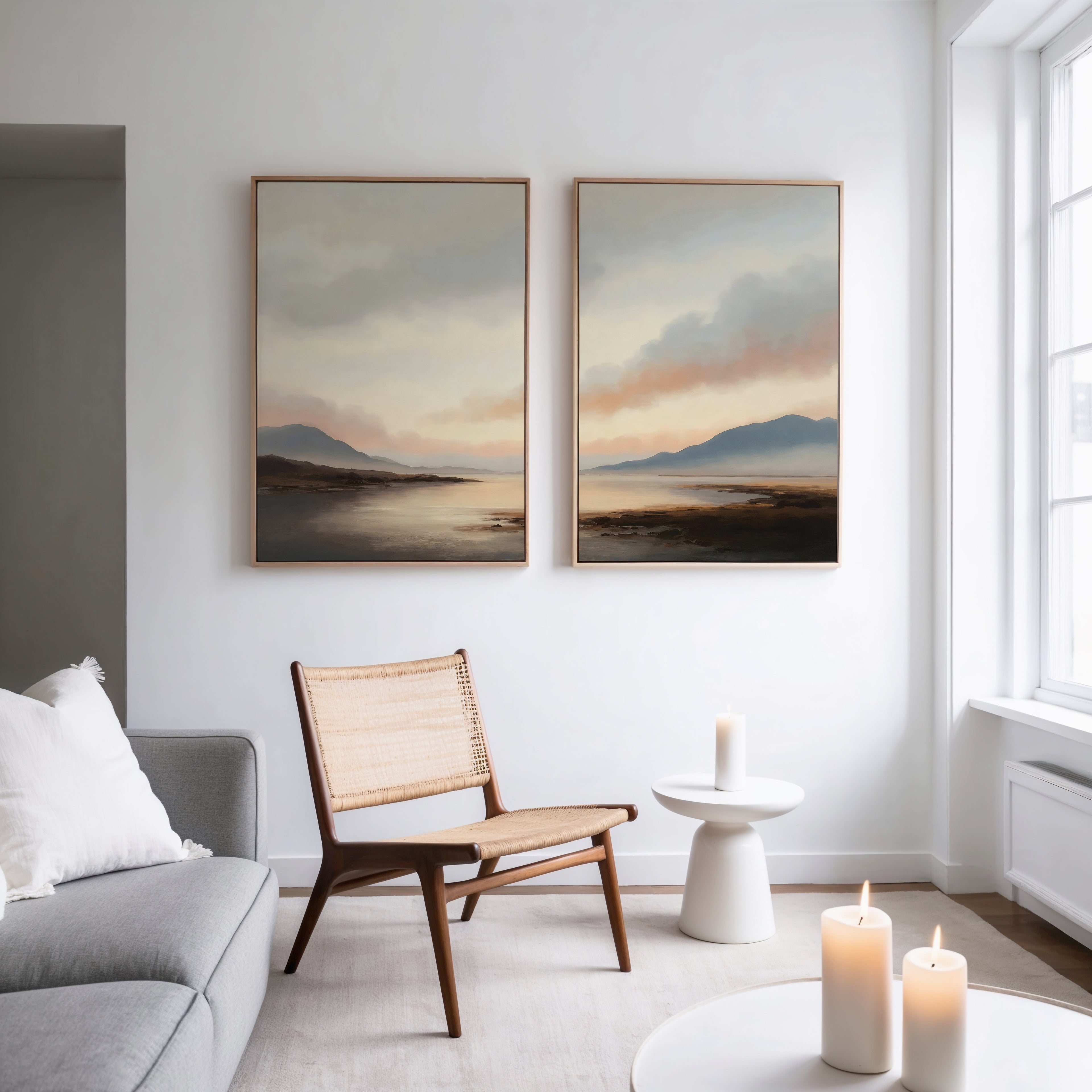 A modern living room features a gray sofa, wooden chair, round white side table, and lit candles. Above, the Distant Peaks Set of 2 minimalist wall art hangs on a white wall near large windows.