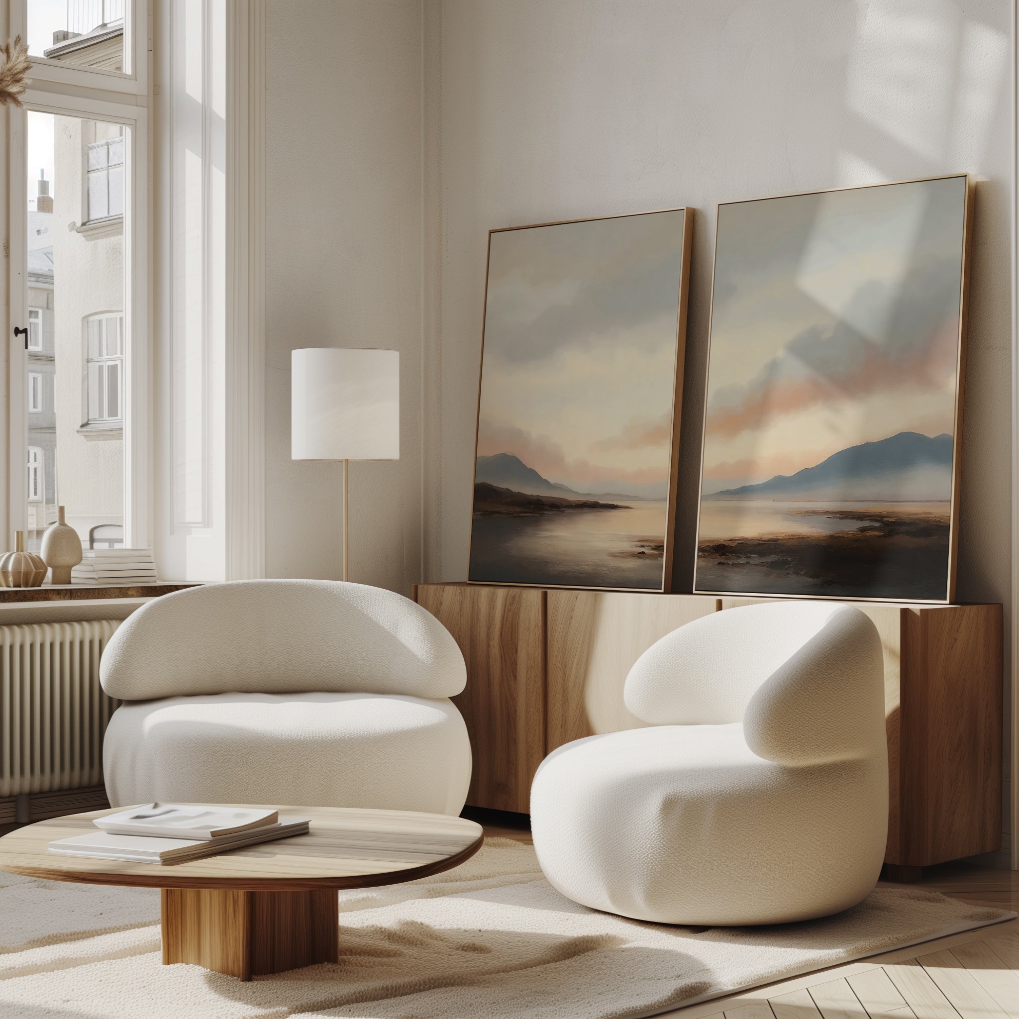 A bright, modern living room features two white armchairs, a low wooden table, and a sideboard adorned with the Distant Peaks Set of 2 wall art. Sunlight streams through tall windows, illuminating the minimalist decor.