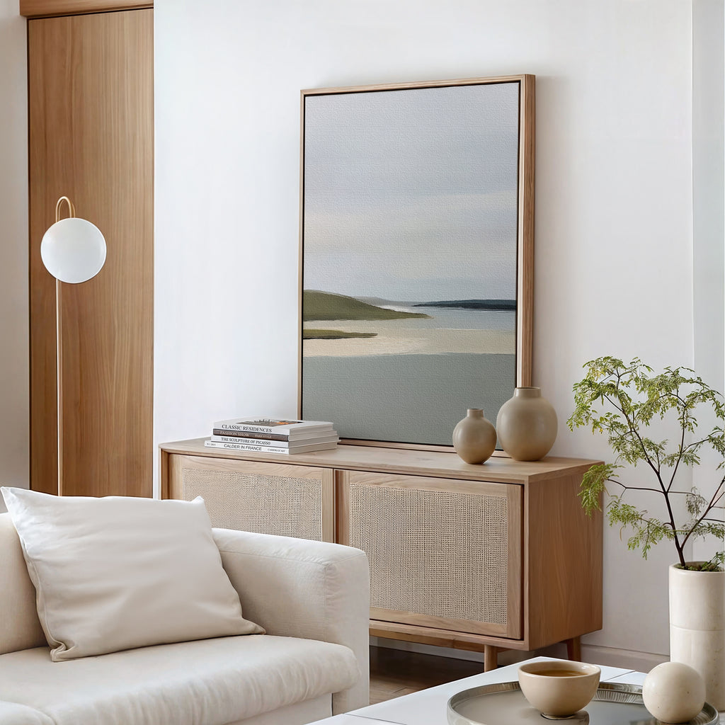 A minimalist living room featuring a light wood sideboard, ceramic vases, books, a Distant Shores Canvas Art as wall decor, a round lamp, a white sofa with cushions, and nature-inspired accents like a small green plant in a vase.