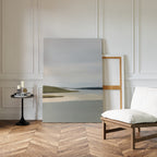 A minimalist room with a patterned wooden floor, white walls, an empty chair with a white cushion, and a black side table holding candles and books. The Distant Shores Canvas Art rests against the wall, adding subtle sophistication.