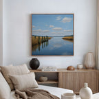 A modern living room with neutral tones displays the Dockside Reflection Canvas Art—showing a serene river scene—hung above a wooden sideboard decorated with vases and books.
