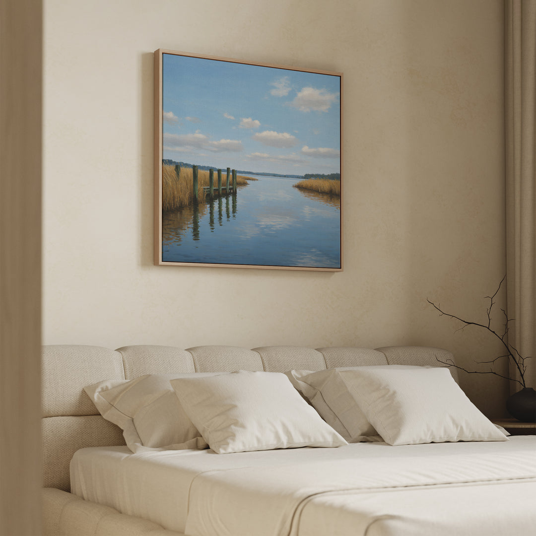 A cozy bedroom features a beige upholstered bed with white pillows and the Dockside Reflection Canvas Art above, displaying a serene lakeside scene with blue sky, clouds, water, and tall grasses.