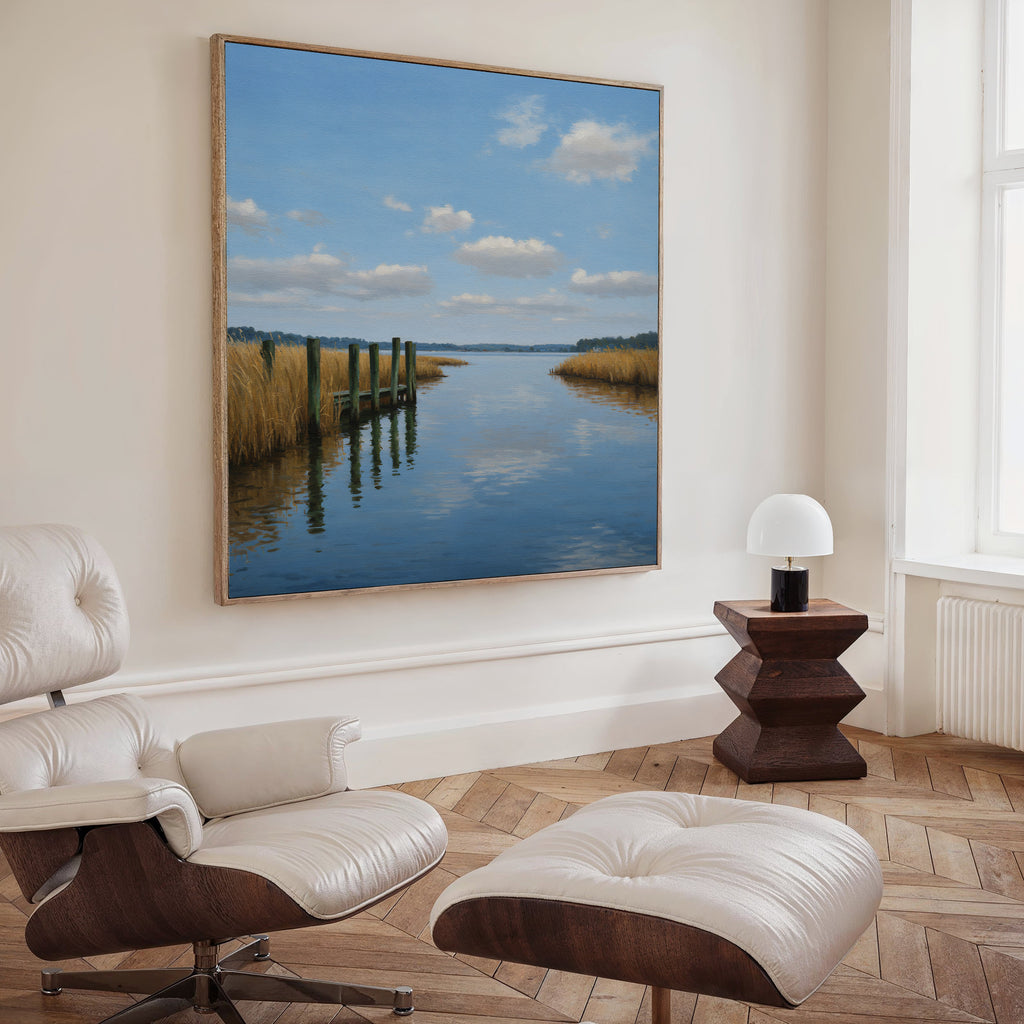 A modern living room features a white leather lounge chair with matching ottoman, dark wood side table with a small white lamp, and the Dockside Reflection Canvas Art depicting a calm river and reeds as striking wall decor.