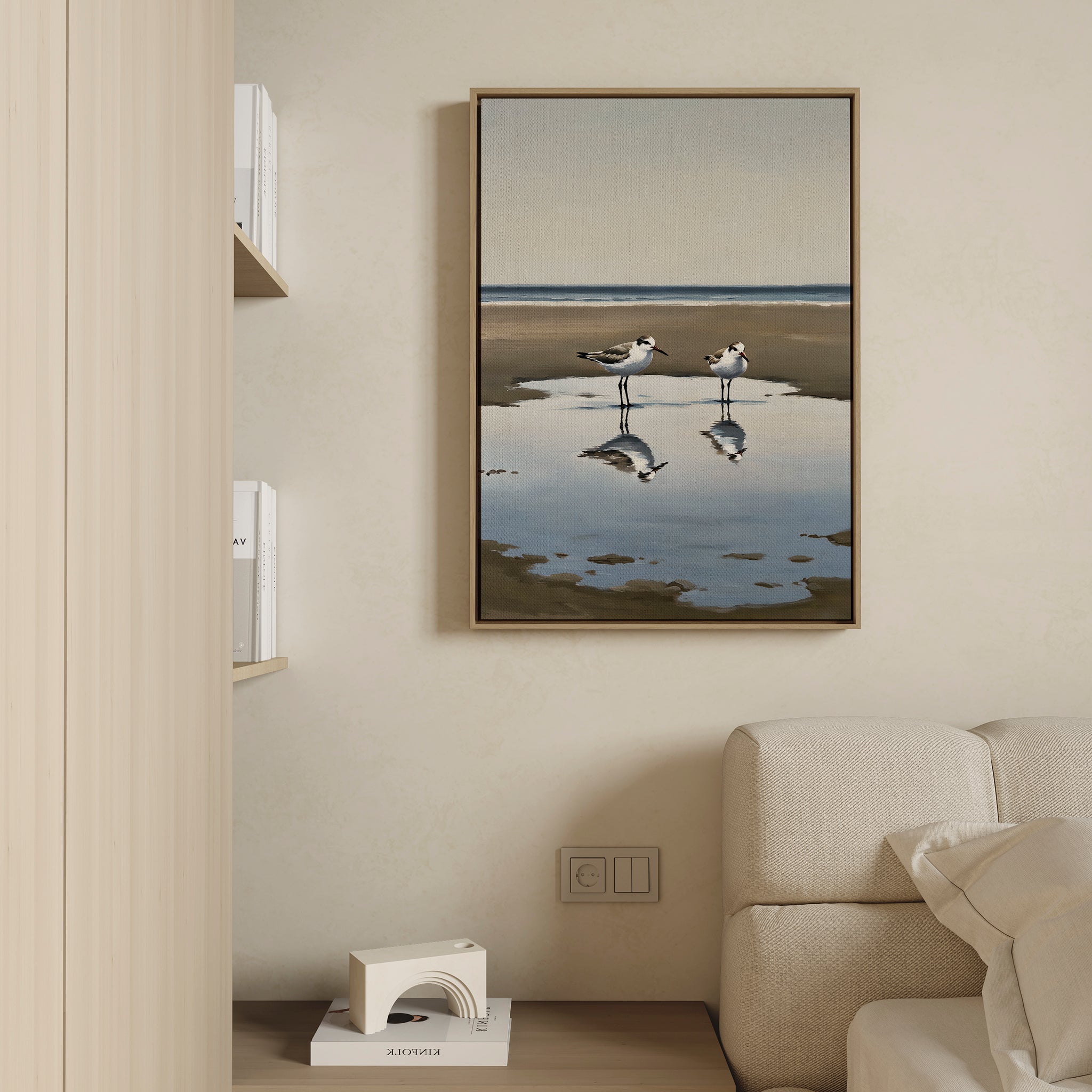 The Drifted Calm Canvas Art, featuring two shorebirds and their reflections, hangs on a beige wall above a light sofa, bringing minimalist style to this contemporary space with shelves displaying books and decor.