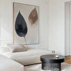 A modern living room with a cream sofa, a round black coffee table with candles and a cup, and the Dual Leaf Study Canvas Art—minimalist wall decor featuring two abstract leaves, one dark and one light, on a white background.