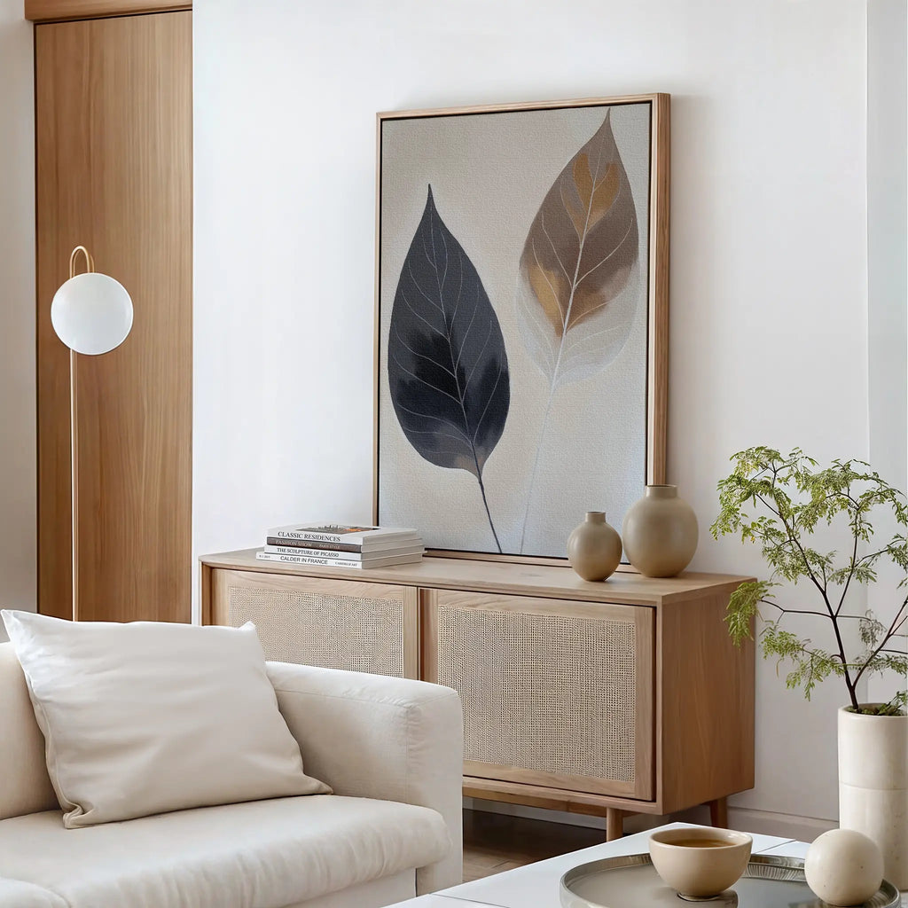 A minimalist living room featuring a light wood sideboard, neutral vases, a stack of books, a potted plant, and a white sofa with a cushion. The serene space is accented by the Dual Leaf Study Canvas Art on the wall.