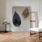 A minimalist room with wooden herringbone floors displays Dual Leaf Study Canvas Art—featuring two contrasting leaves—resting against the wall. Nearby sit a small black side table with candles and books, plus a white cushioned chair.
