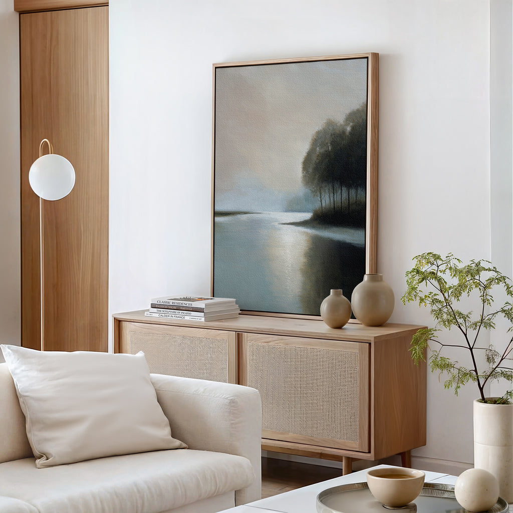 A modern living room with neutral tones features a light wooden sideboard, calming decor, ceramic accents, books, a white cushion sofa, a potted plant, a round hanging lamp, and Dusklake Canvas Art as large framed wall art of trees by water.