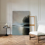 Dusklake Canvas Art, a large minimalist landscape painting of a lake and trees, rests against a white paneled wall in a stylish room beside a blank wooden frame, cushioned chair, and round table with candles and books.