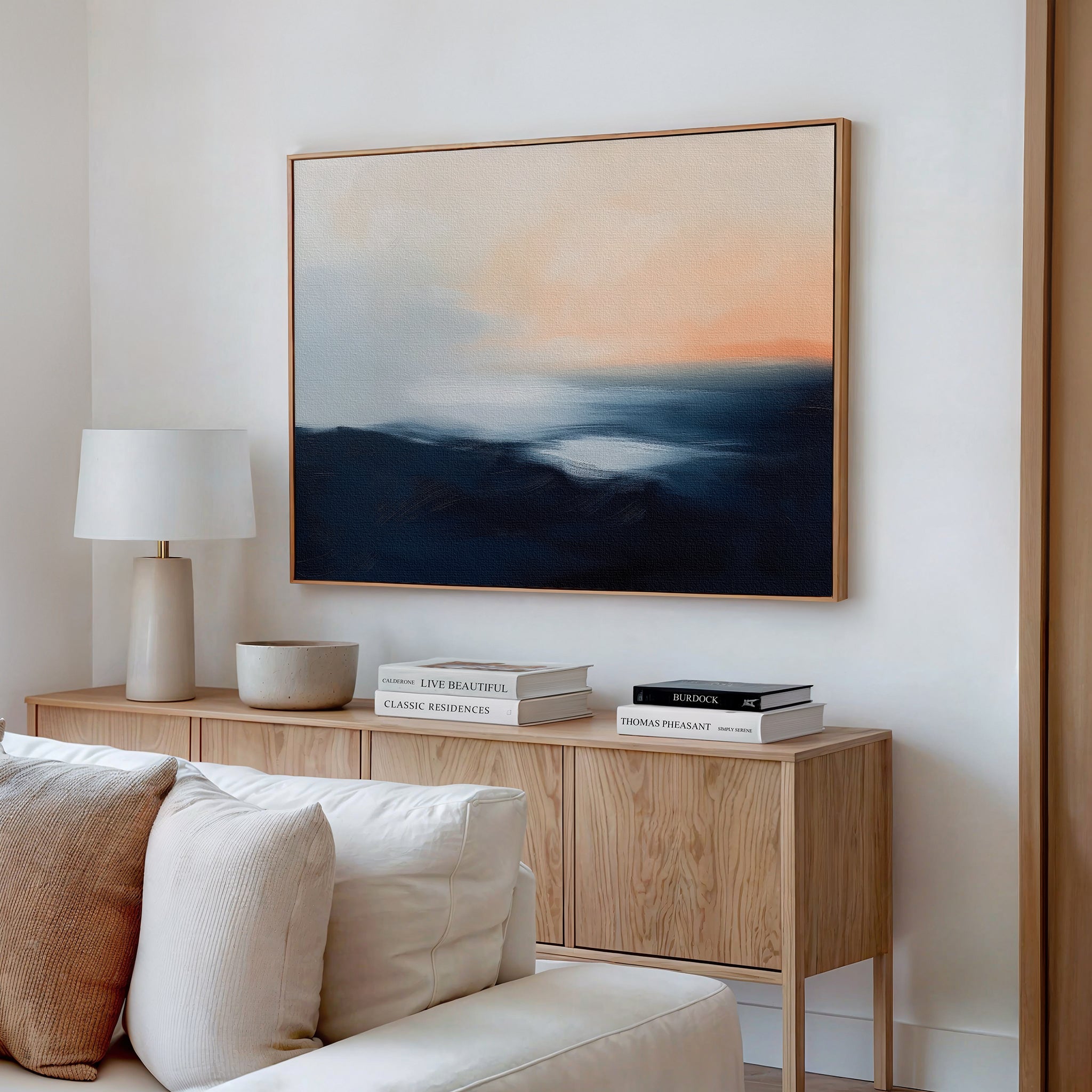 A modern living room features a wooden sideboard with stacked books, a lamp, and a bowl. Above hangs the Dusk Ridge Canvas Art—a large abstract sunset painting in blue and orange hues. A beige couch is partly seen in the foreground.